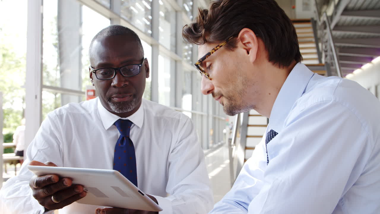 Two Businessmen Looking At Digital Tablet During Meeting Around Table In Modern Office