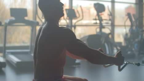 athletic shirtless male doing workouts on a back with power exercise machine in a gym club. side view of muscular man which using rowing machine in gym