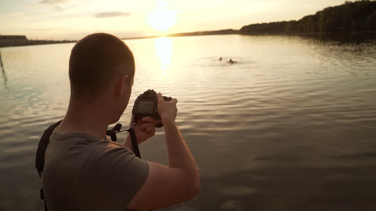 A man is standing with his back and photographing the sunset on the lake surrounded by trees on the background with bathing people.