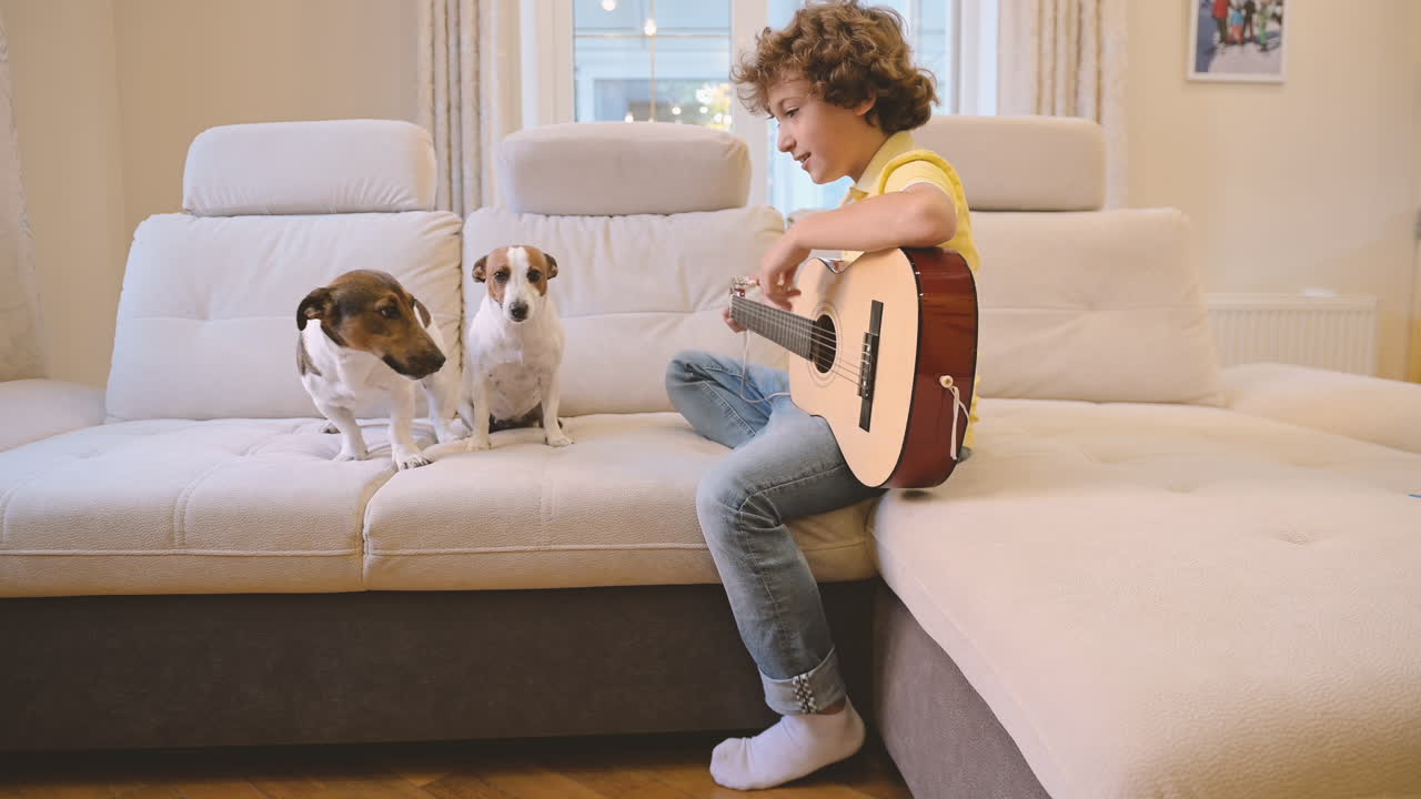 chico rubio con el pelo rizado tocando la guitarra sentado en el sofá, junto a él están sus perros tumbados y caminando en el sofá