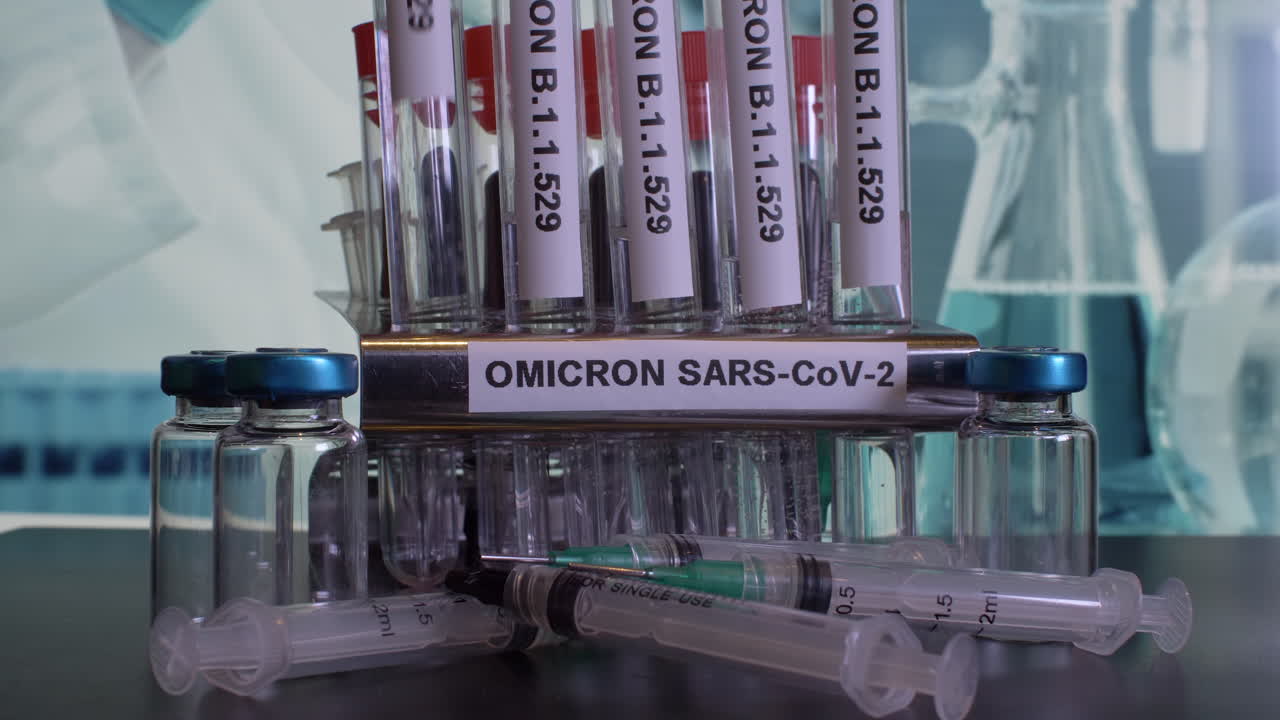 A frontal look on a  metal tube rack labeled OMICRON SARS-CoV-2. The glass test tubes named B.1.1.529 are put into the rack. A few syringes lie in front of a rack. The laboratory background is blurry