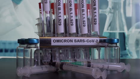 a frontal look on a  metal tube rack labeled omicron sars-cov-2. the glass test tubes named b.1.1.529 are put into the rack. a few syringes lie in front of a rack. the laboratory background is blurry