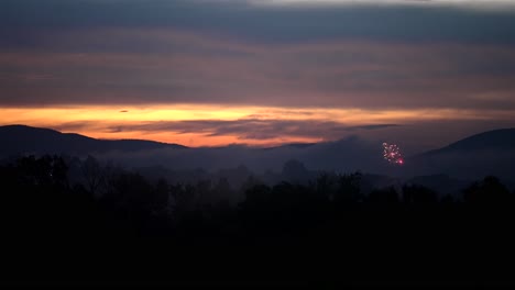 Overview-of-fireworks-launching-into-air-at-dusk-with-orange-red-glow-on-horizon-below-mountain-silhouette