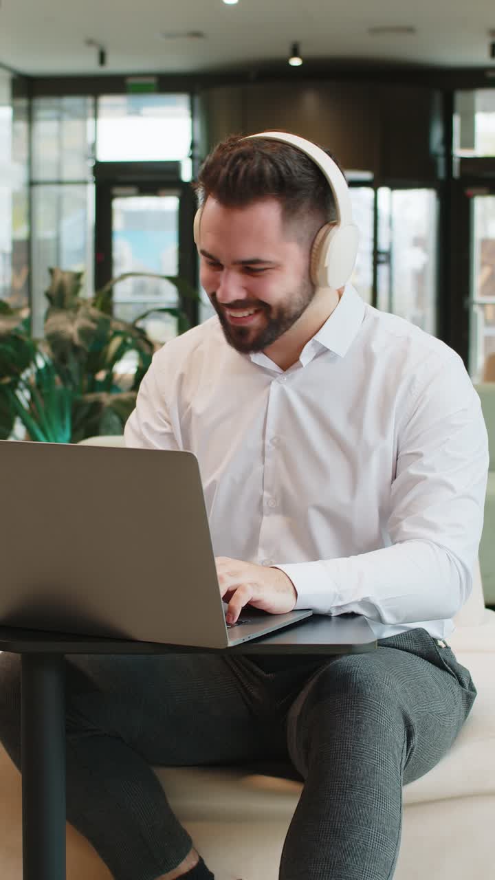 joven empresario sonriente usando una computadora portátil y escuchando música a través de auriculares en el vestíbulo de una oficina moderna