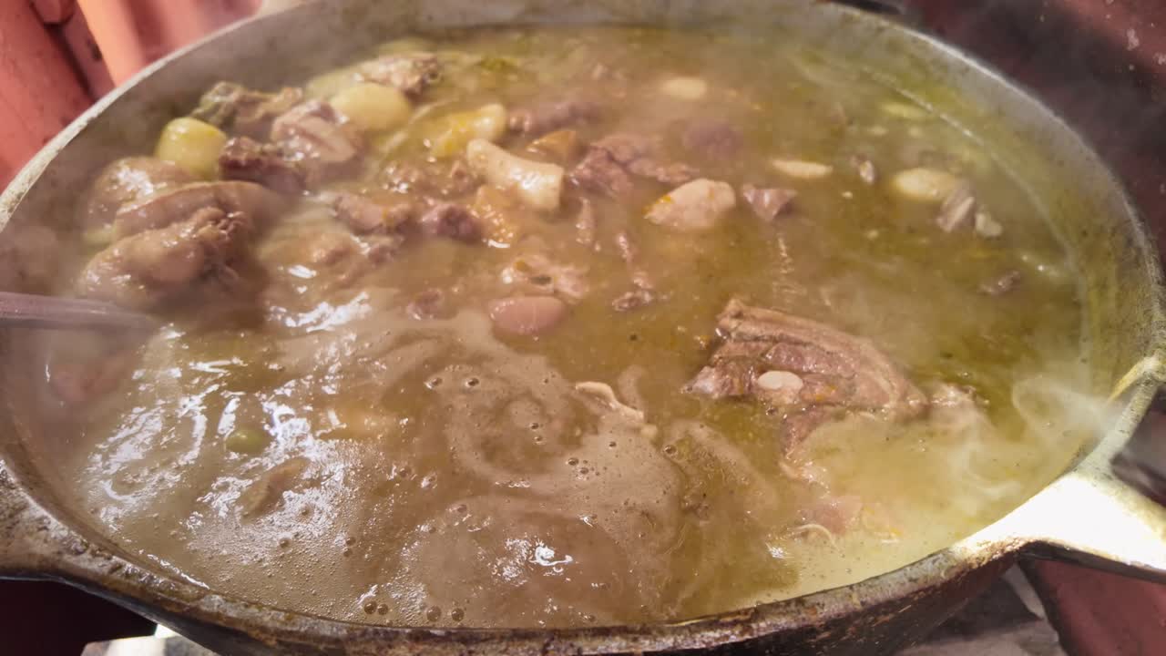 Vista em close-up de uma panela de metal cheia de Sancocho Dominicano cozinhando, enquanto uma colher mexe os ingredientes, e o vapor envolve as carnes e vegetais neste prato tradicional