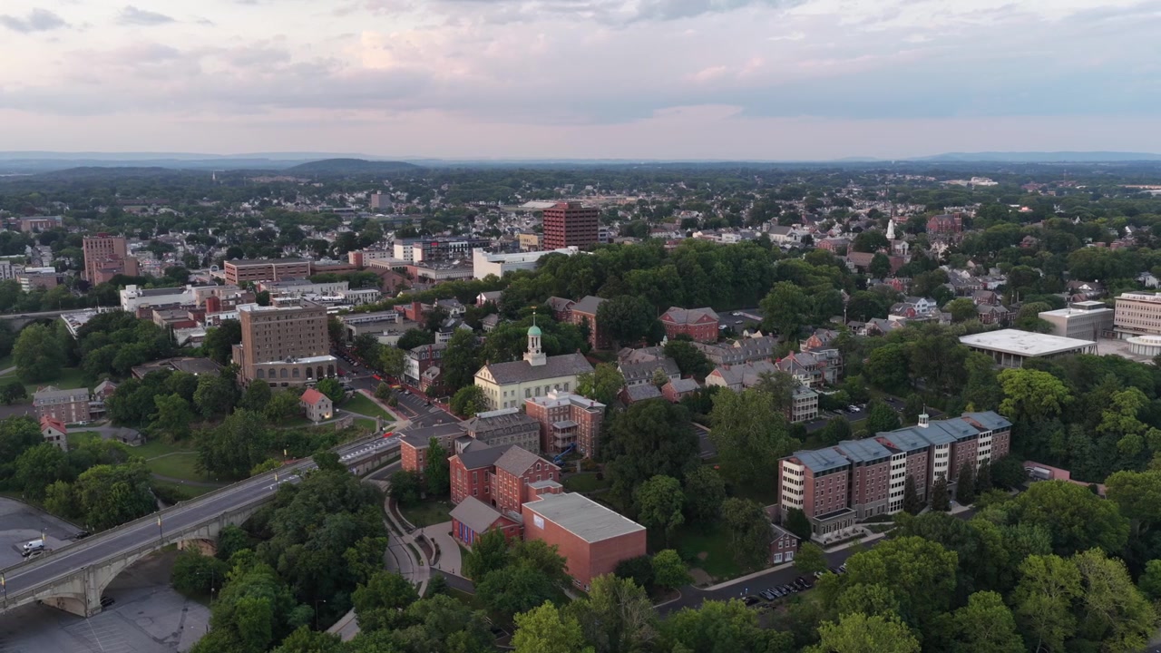 Case storiche con tetti a capanna ripidi fanno capolino attraverso la chioma degli alberi estivi, lo skyline del centro di Bethlehem si erge oltre le colline boscose in un quartiere residenziale della Pennsylvania. Ampia ripresa aerea da drone.