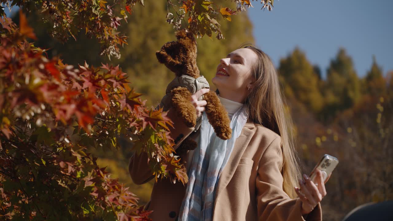 Woman Taking Selfie with Mobile Phone Holding Brown Toy Poodle Puppy In her Arms Standing By Colorful Maple Tree Foliage in Autumn Park - slow motion