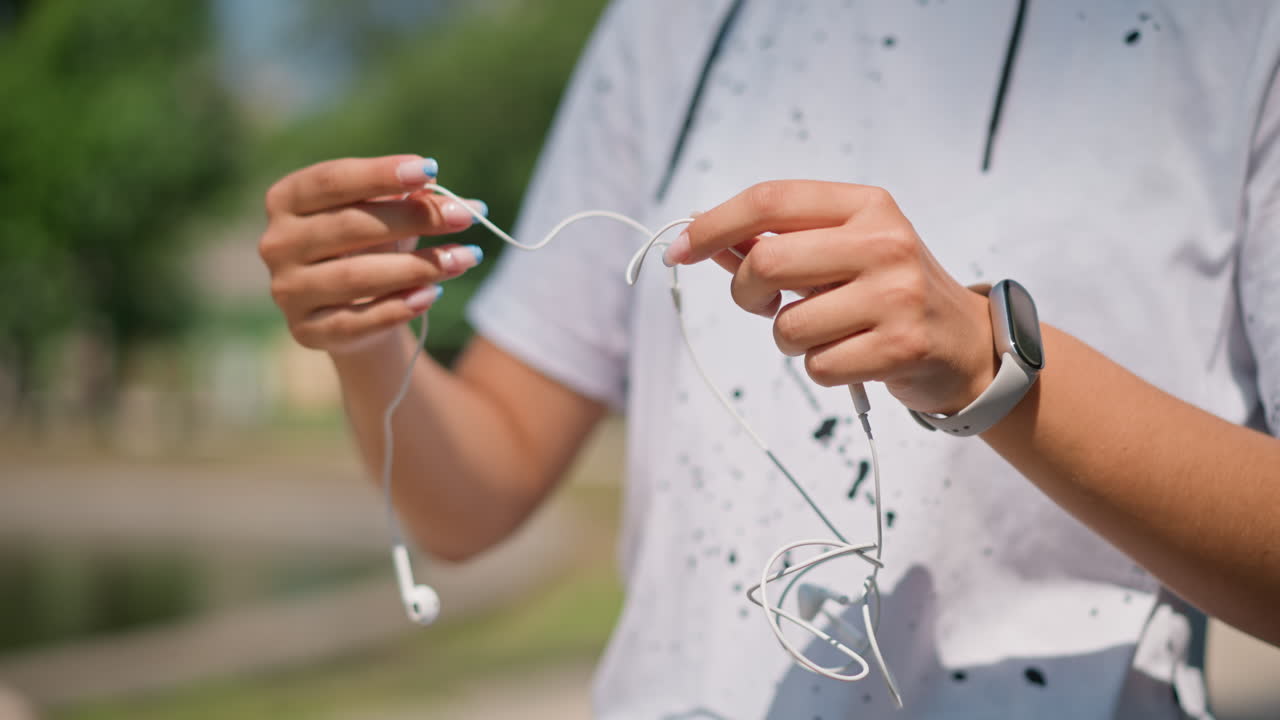 공원 산책로 근처에서 엉킨 이어폰을 풀고 있는 음악가, 매듭진 케이블을 정리하는 조심스러운 손길, 보이는 스마트워치, 리허설을 위한 집중된 준비, 자연광과 차분한 분위기