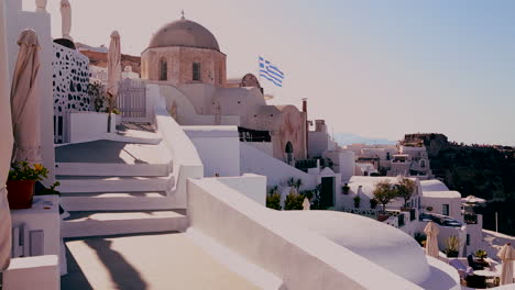 white houses line the hillsides of the greek island of santorini with a greek flag in the distance