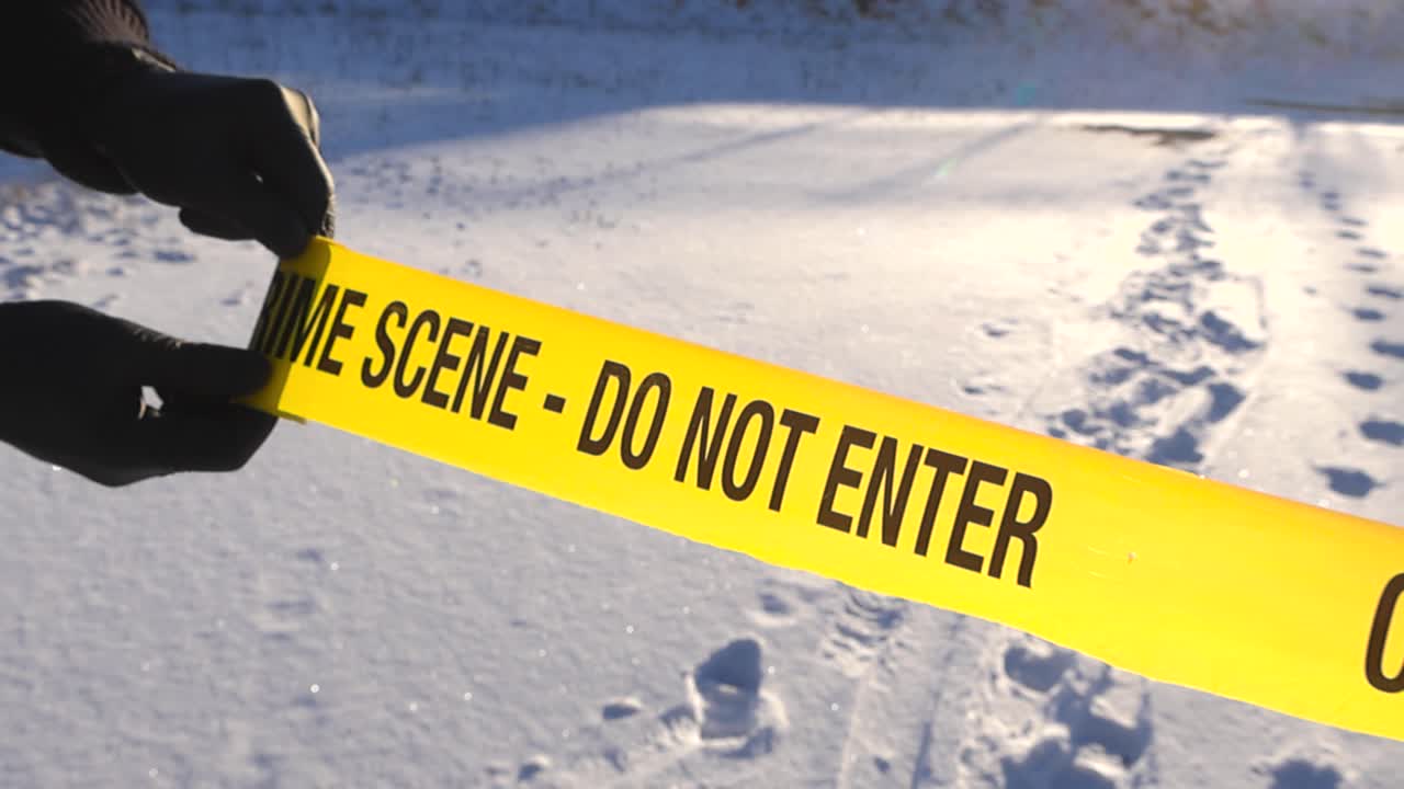 Police forensic science investigator unrolling and closing off a winter white snow covered area with a crime scene yellow colored tape ribbon at day. Sunshine backlits the tape making it glow