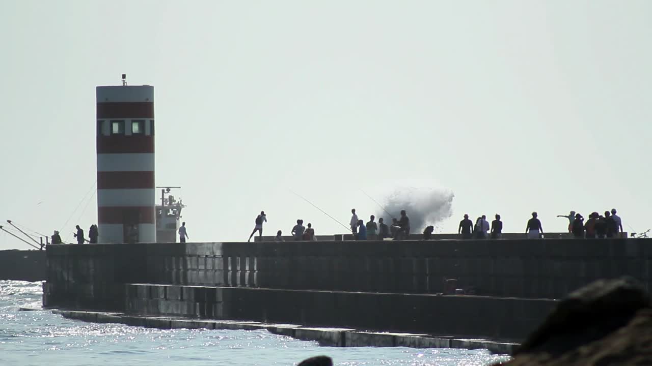 Silhouette of a lighthouse at a harbor getting hit by big waves while tourists are walking along.