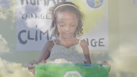 animation of clouds over smiling biracial schoolgirl holding recycling box in ecology class