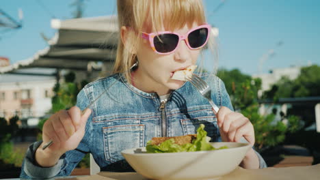 a girl in pink glasses is eating a salad on the summer playground of a cafe summer holiday with kids