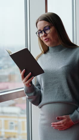 positive smiling pregnant woman wearing glasses reads a book. lady stands at the window touching her belly. vertical video