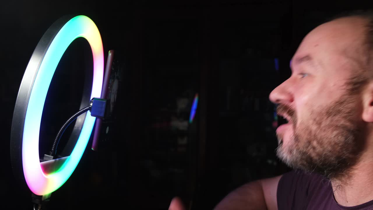 면도되지 않은 남자가 반지 램프와 드폰 앞에 앉아 vlog을 하고, 승인하는 손짓을 보여주는 비디오를 녹음합니다.