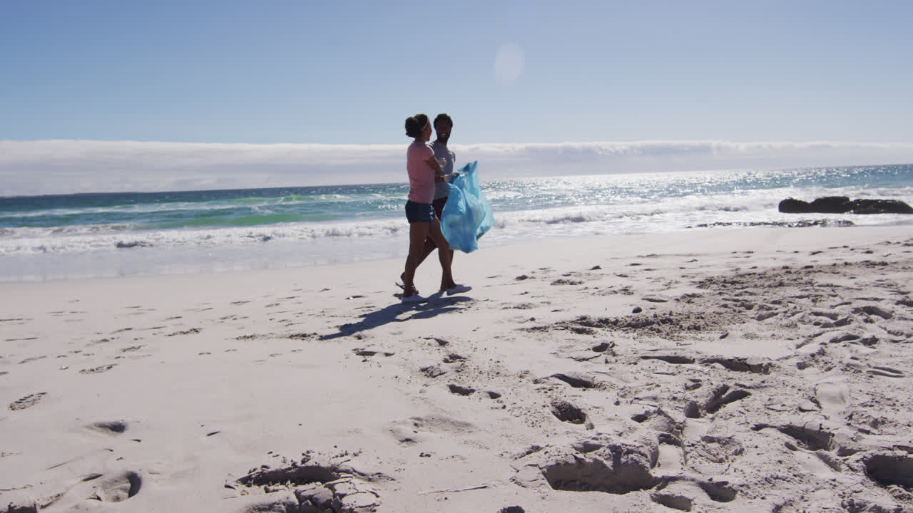 una pareja afroamericana caminando con bolsas de basura y recogiendo basura de la playa