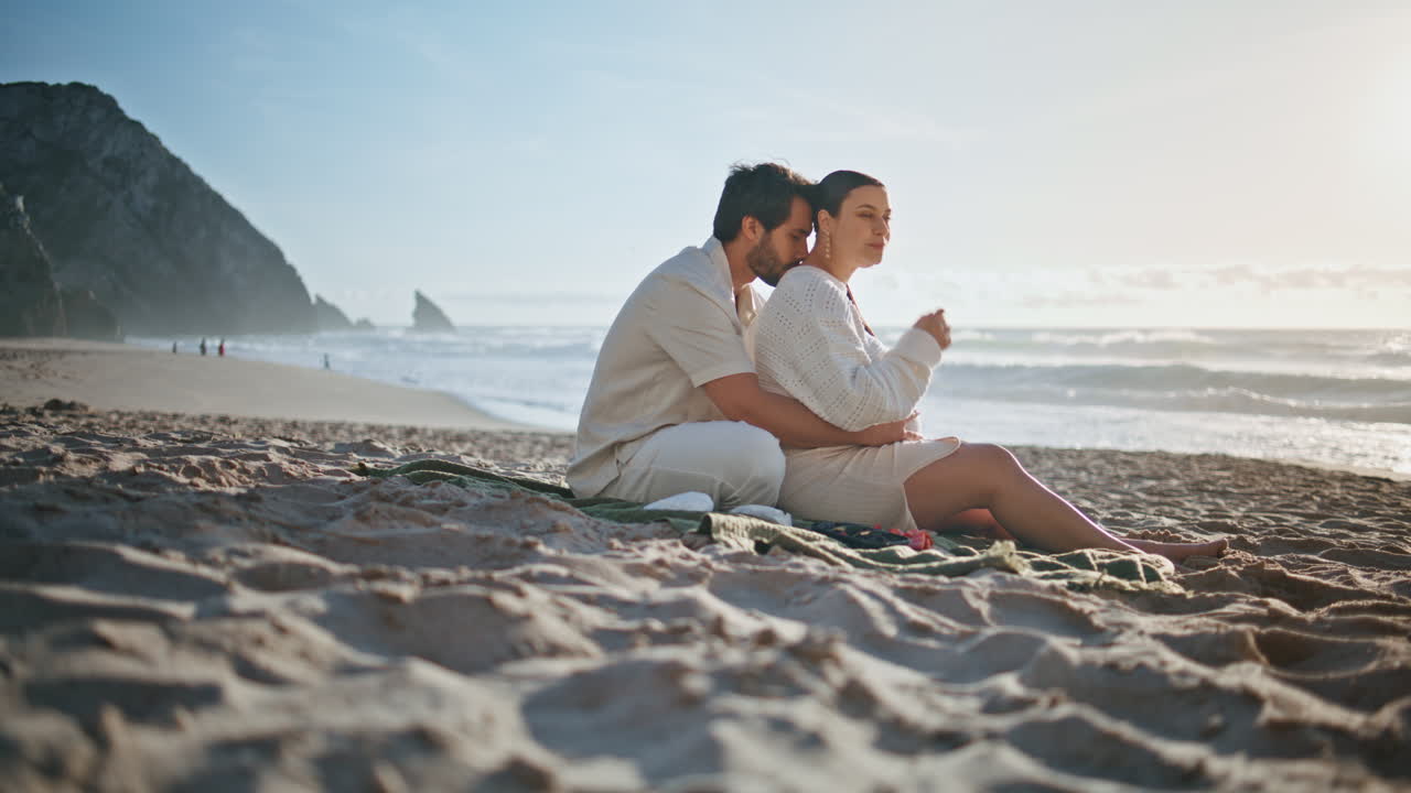 임신 한 커플이 모래 해변에서 휴식을 취하고 있습니다. 바다를 바라보는 커플이 아기를 기대하고 있습니다.