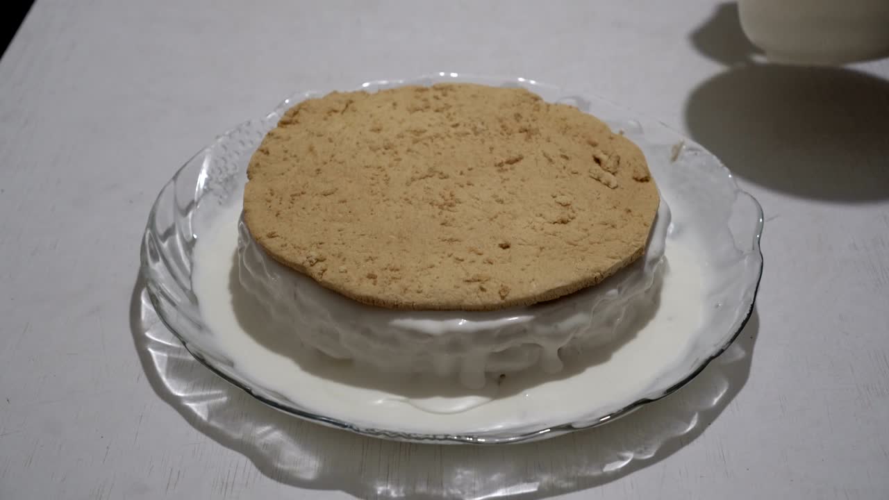 A female chef prepares a layered biscuit cake and lubricates it with cream and white chocolate. Cake on a glass plate. Preparation of sweet dessert. Close-up. The view from the top. 4K.