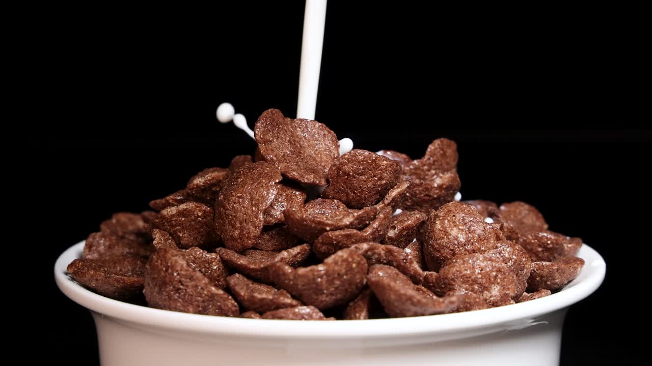 Milk cascades over crunchy chocolate cereal in a white bowl against a black background, creating a dynamic splash effect