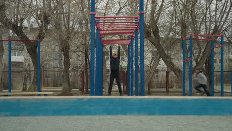 a coach swings on a red iron bar in an outdoor fitness area, with people playing football in the background, surrounded by bare trees and residential buildings