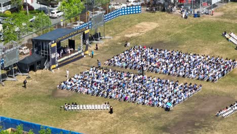 Aerial-view-showing-high-school-graduation-outdoors-on-grass-field-with-stage-during-sunny-day---I