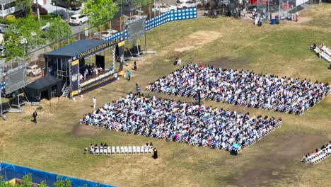 aerial view showing high school graduation outdoors on grass field with stage during sunny day - i