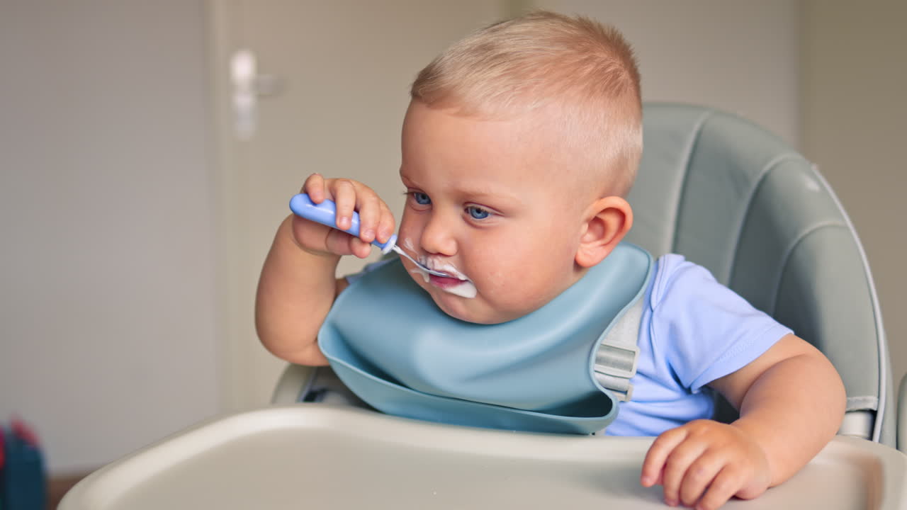 Toddler boy eats independently using a baby spoon. One and a half year old boy child eats yogurt. The toddler finishes eating and holds it out to the camera