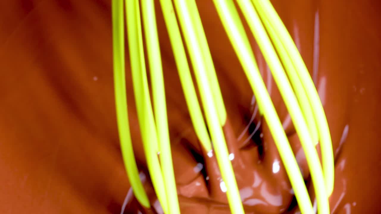 A bright green whisk stirring smooth, melted chocolate, creating swirls and textures in the rich, glossy mixture.