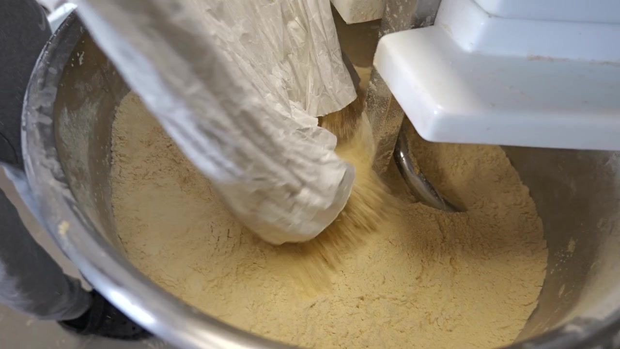 Overhead view of a worker emptying a bag of flour into a large industrial mixer at the beginning of the fresh pasta making process.