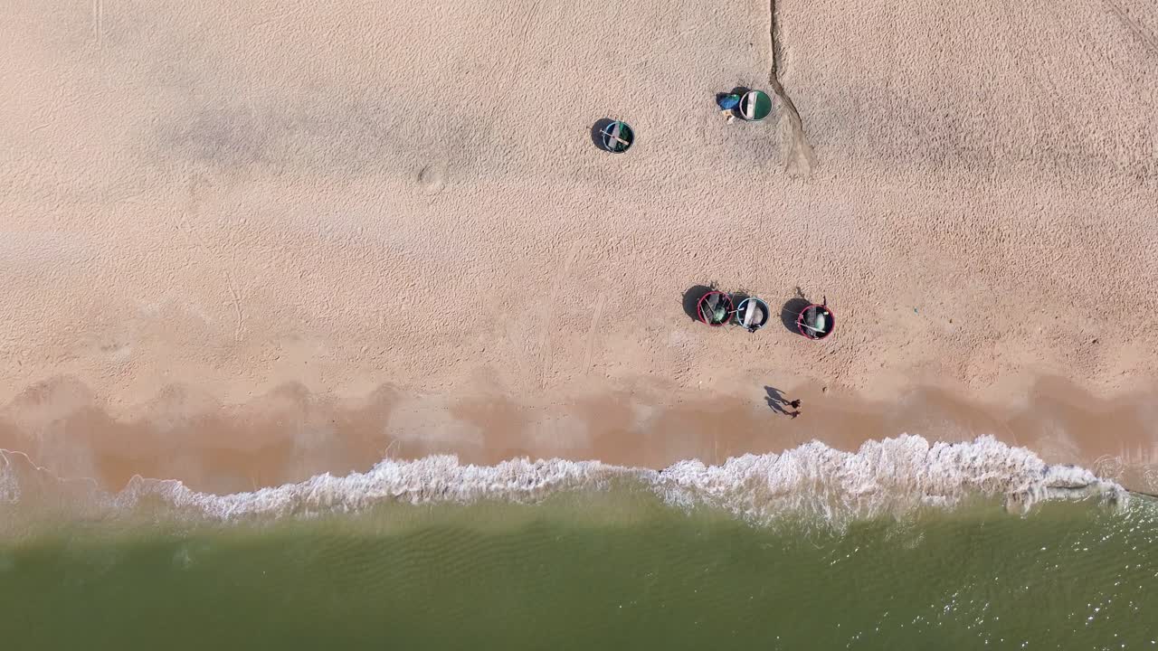 Drone hovers over Mui Ne shore in bird’s-eye view, filming people walking along the beach and bucket boats parked close on the pristine white sand.
