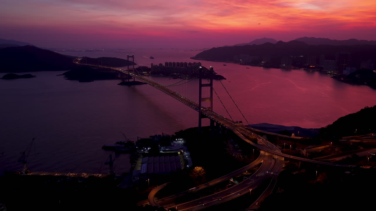 Cinematic Aerial view of Tsing Ma Bridge in Hong Kong at dusk, illuminated against a vibrant pink and purple sunset sky, spanning tranquil waters with city skyline and mountains in the background