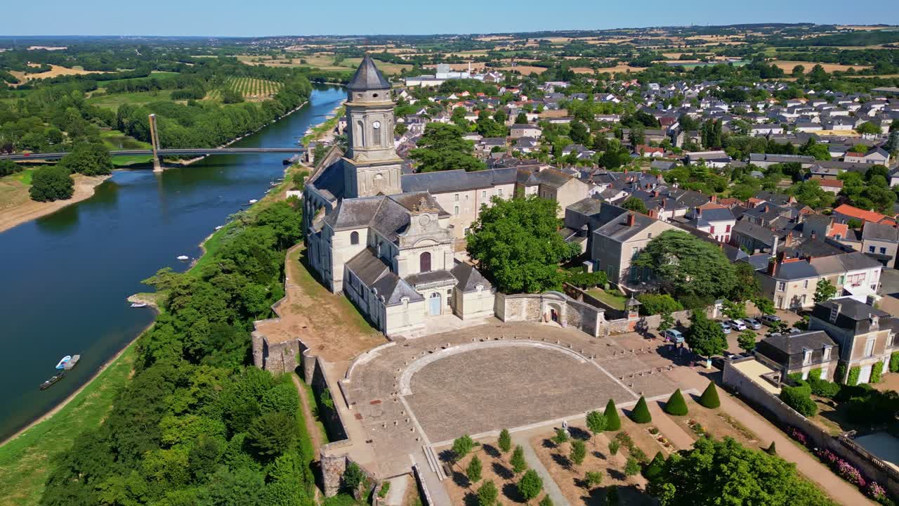 Drone side panning shot from Loire River over trees to Saint-Florent Abbey Church, showing garden, square, houses, bridge, fields, and vegetation - Mont Glonne in France