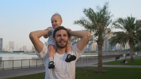 Un-Padre-Amoroso-Con-Una-Camiseta-Blanca-En-El-Verano-Camina-Con-Un-Niño-Sentado-En-El-Cuello-Contra-La-Ciudad-Y-Las-Palmeras