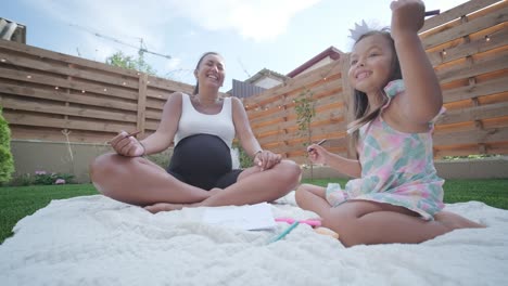 una madre y su hija pequeña se sientan en una manta en el patio trasero, participando en una actividad de dibujo juntos. la escena al aire libre captura un momento familiar pacífico, fomentando la creatividad y el vínculo.
