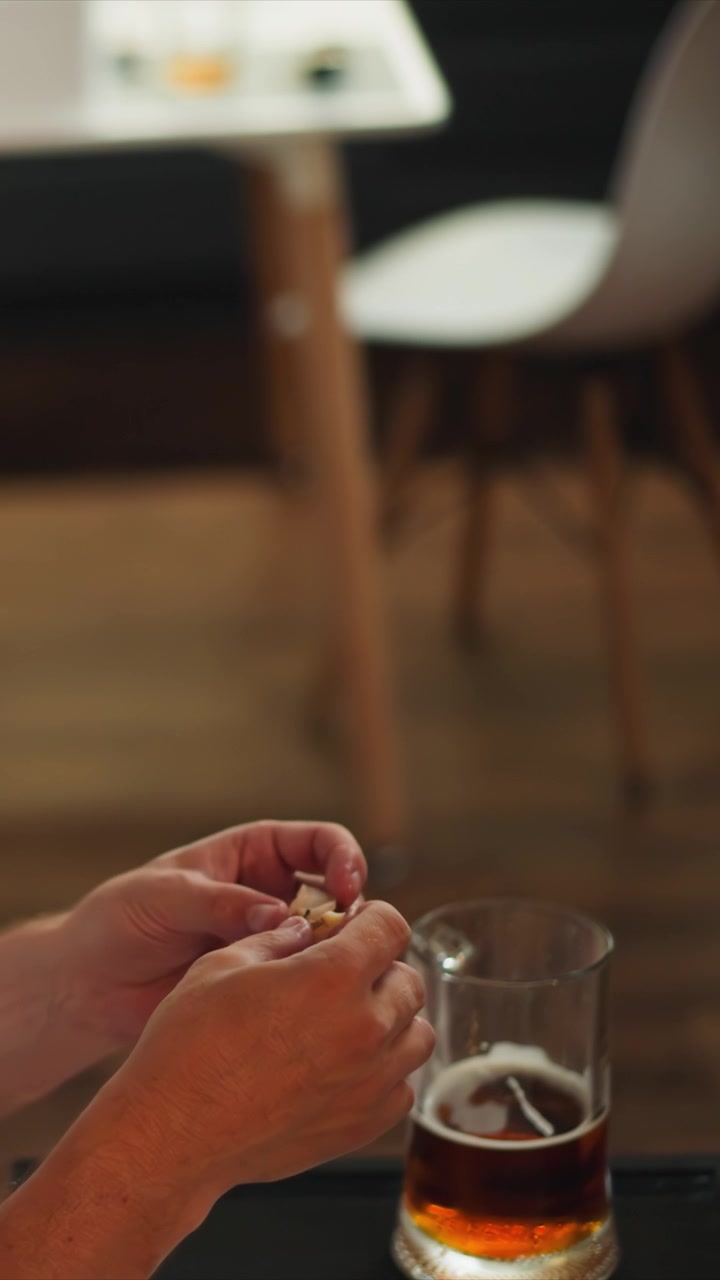 Primo piano di mani che mangiano uno snack con birra in un bicchiere sul tavolo. Le mani manipolano l'alimento mentre la bevanda parzialmente visibile rimane ferma lì vicino. Ambiente interno con sfondo sfocato e un'atmosfera calda.