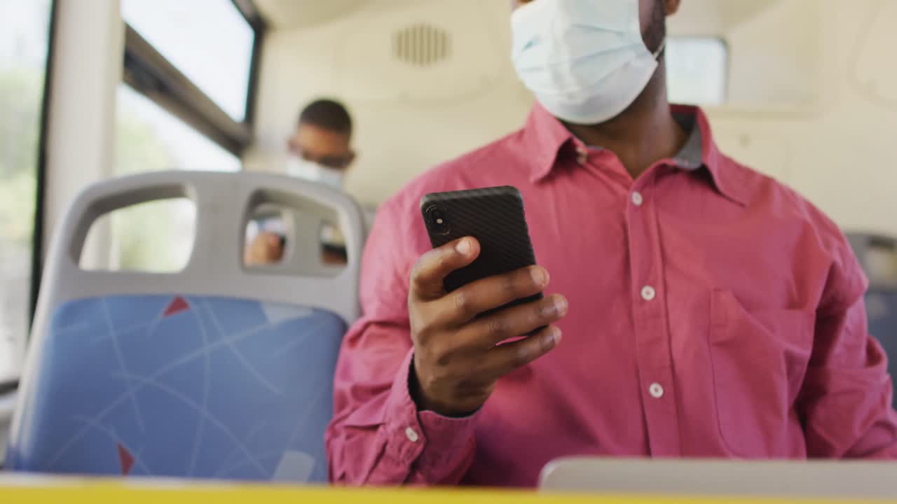 sección media de un hombre afroamericano en la ciudad con una máscara facial, usando un teléfono inteligente en el autobús