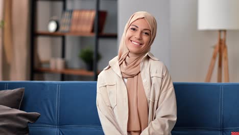 portrait of smiling young islamic woman in hijab middle eastern muslim lady sitting on sofa at home