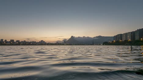 Low-to-the-water-time-lapse-of-sunset-at-city-lake-in-Rio-de-Janeiro-setting-behind-the-Two-Brothers-mountain-veiled-in-clouds-and-floating-Christmas-tree-turning-lights-on