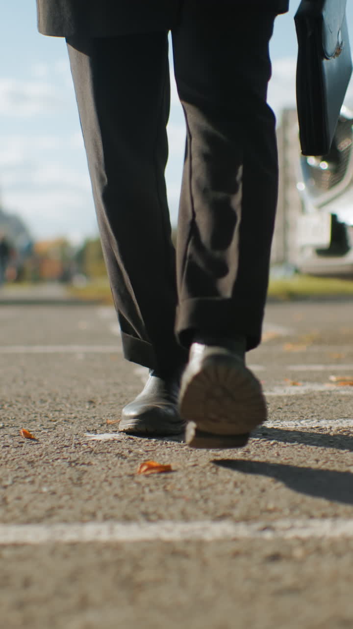 vista de la pierna del hombre de negro caminando con confianza más allá de los coches estacionados en el pavimento urbano bajo la luz del sol, llevando un maletín, rodeado de hojas de otoño, con vista borrosa de personas caminando en el fondo