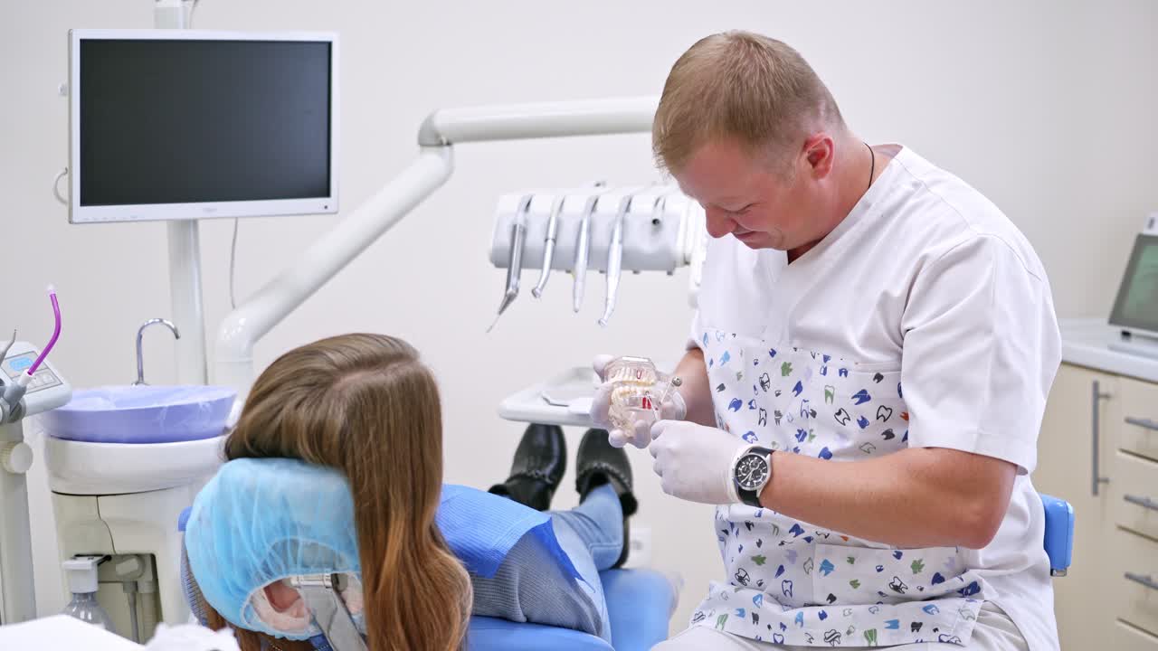 Doctor shows his young blonde woman patient on a plastic jaw sample how he will treat her teeth. Modern dental clinic background. Side view video. Cheerful stomatologist.