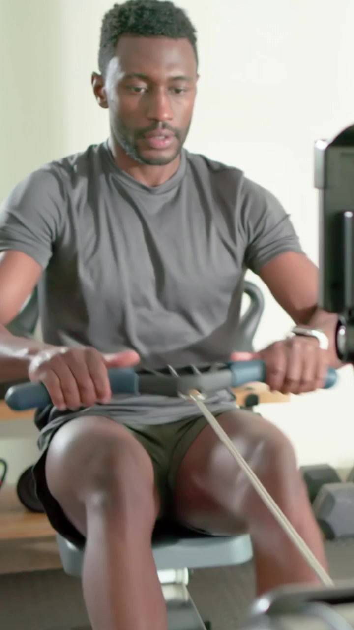 Vertical video: Rowing man in gray shirt, olive shorts leaning forward driving on rower for cardio