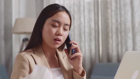 close up of asian businesswoman holding a thermometer and talking on phone while working with laptop in the living room at home