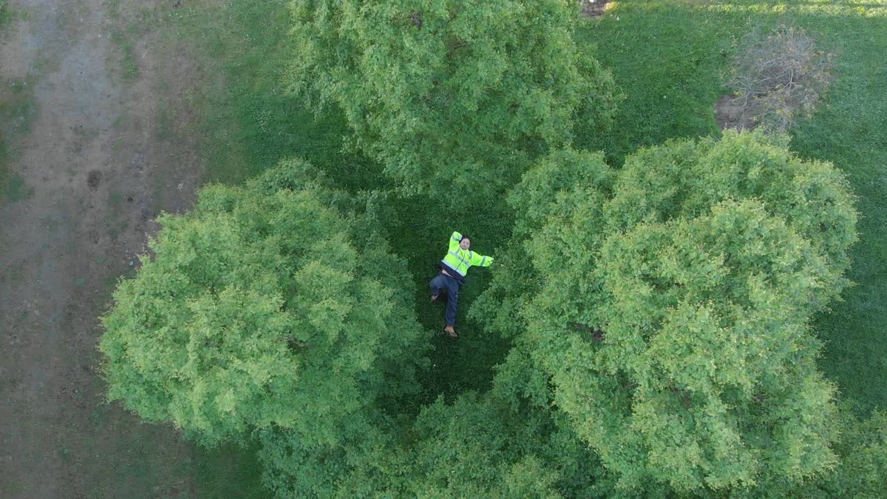 A warehouse worker taking a break, laying on the green grass, relaxing - aerial ascend