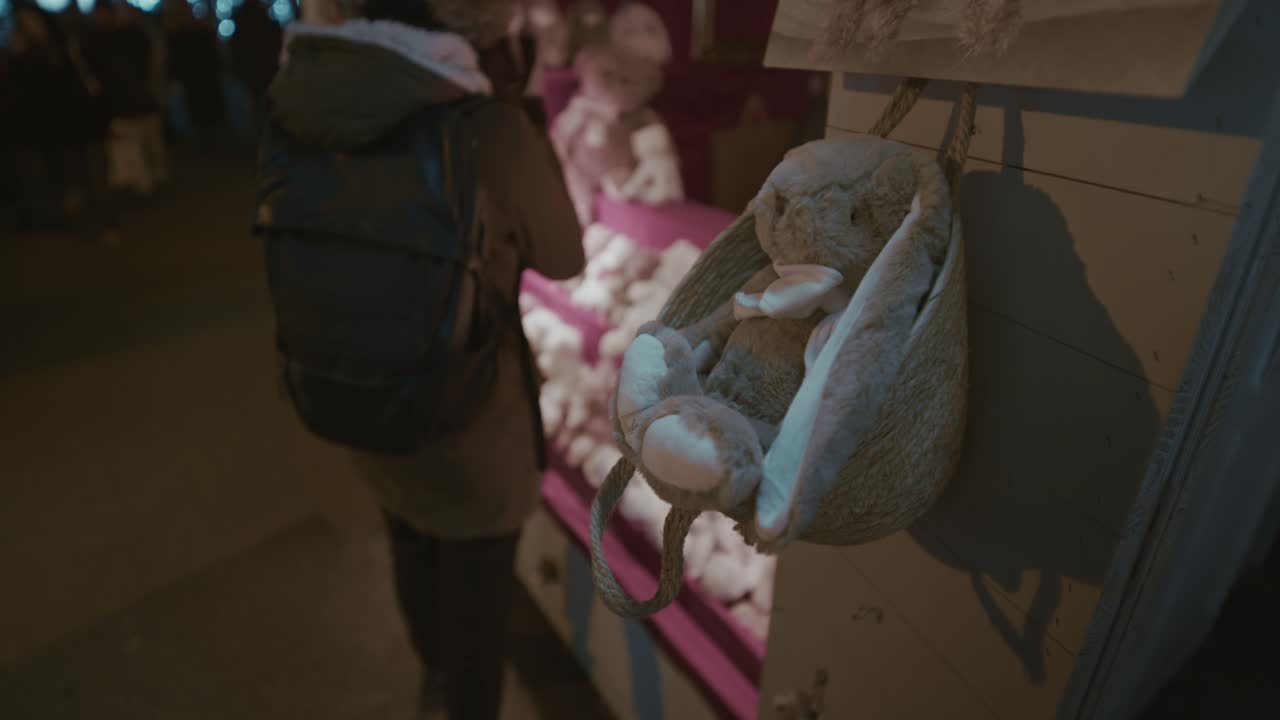 Un coniglietto di peluche appeso a una corda su un banco del mercato, con altri peluche morbidi esposti sullo sfondo durante una serata invernale
