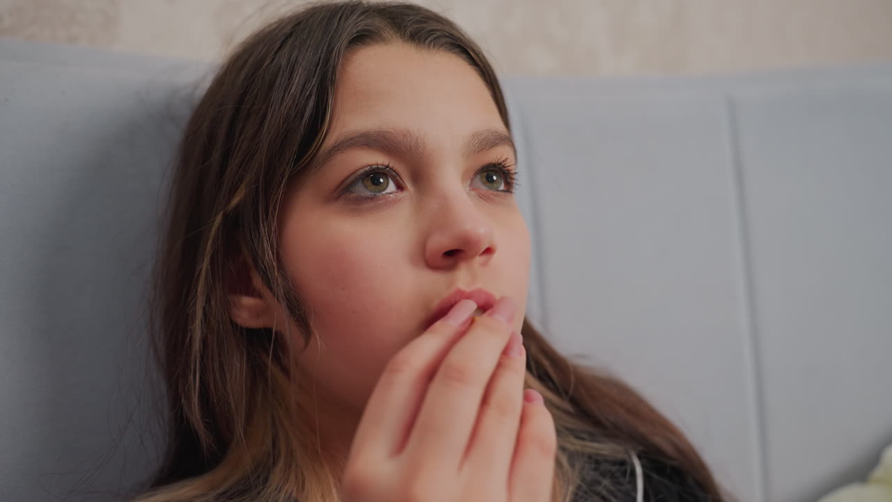 Close up of brunette girl holding popcorn near lips while watching something off screen, girl sits on bed with calm expression and focused gaze, suggesting she is watching movie