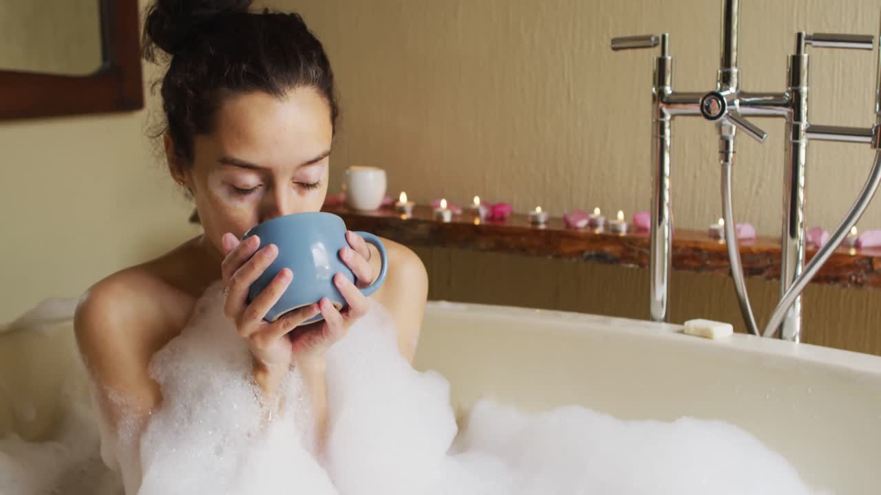 Relaxed biracial woman lying in bath with foam and drinking coffee