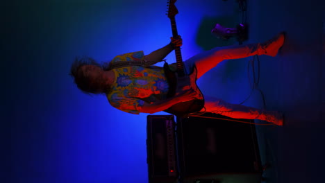 vertical video of a crazy jumping rock guitarist with a guitar in his hands on a colored neon background dancing and playing the guitar. energetic guitarist groovy dancing and looking at the camera
