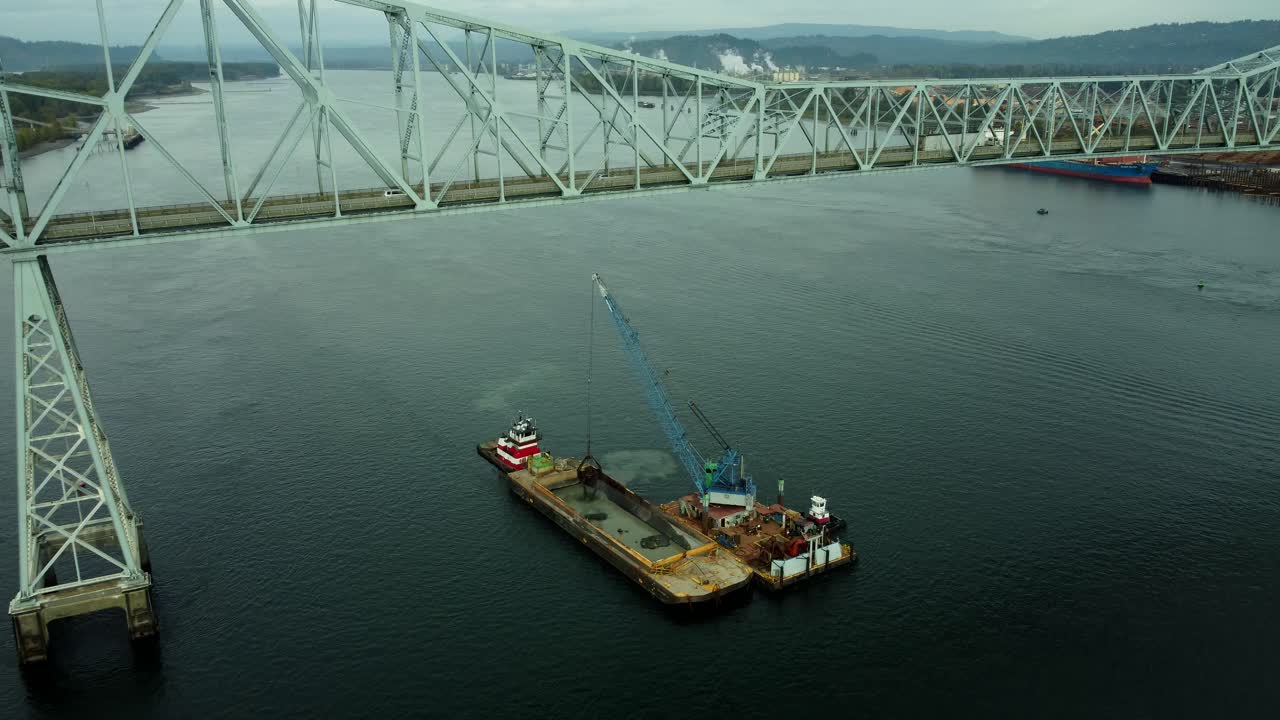 US, WA, Longview, 2025-10-22 - Drone view of the Lewis and Clark Bridge between Oregon and Washington with a sand dredge emptying its bucket into a barge
