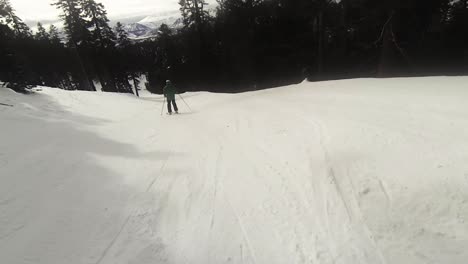 Punto-De-Vista-De-Un-Snowboarder-Persiguiendo-A-Un-Esquiador-Con-Una-Chaqueta-Verde-Bajando-Por-Un-Sendero-Angosto