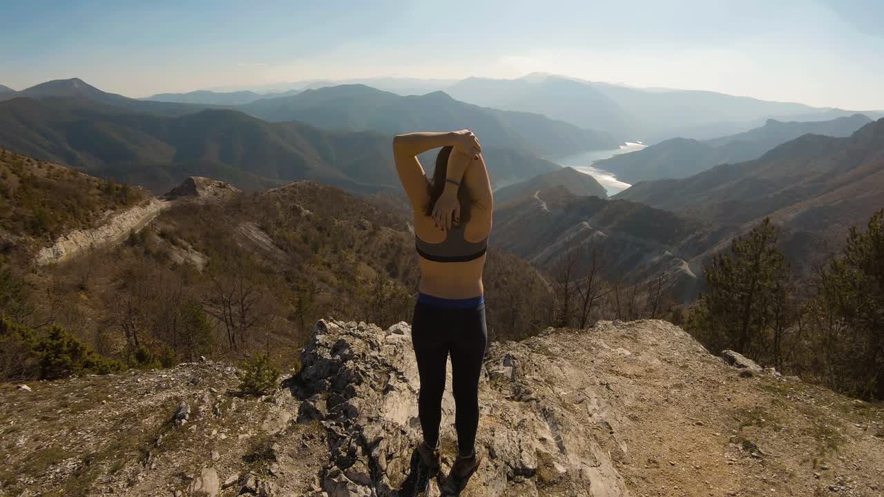 una atleta estirándose en la cima de una montaña con un hermoso lago en el cañón frente a ella.